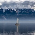 A lone sailboat navigates calm waters against a backdrop of majestic, cloudy mountains.