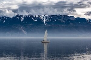 A lone sailboat navigates calm waters against a backdrop of majestic, cloudy mountains.