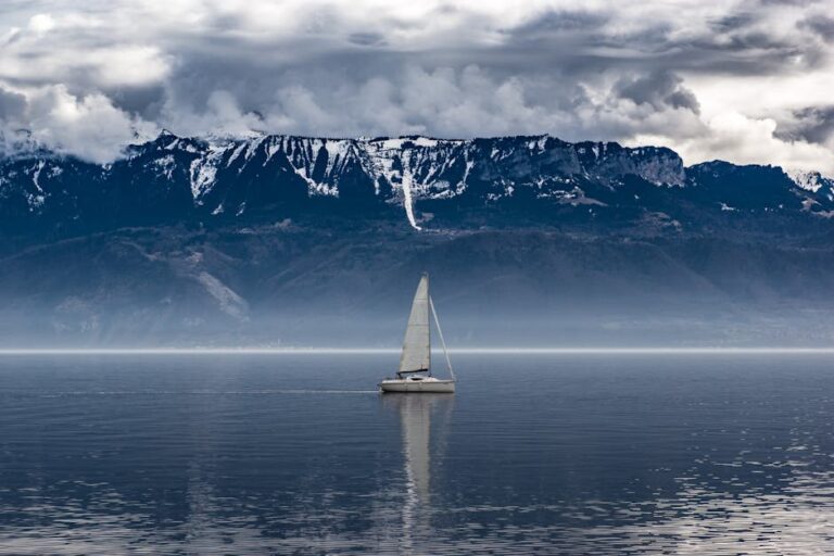 A lone sailboat navigates calm waters against a backdrop of majestic, cloudy mountains.