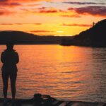 A person stands on a dock, silhouetted against a vibrant sunrise over a tranquil lake.
