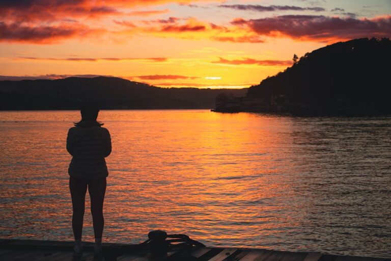 A person stands on a dock, silhouetted against a vibrant sunrise over a tranquil lake.