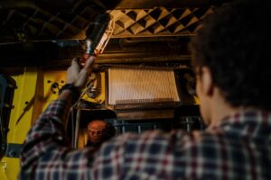 Mechanic inspecting vehicle engine under hood with handheld light, indoors workshop.