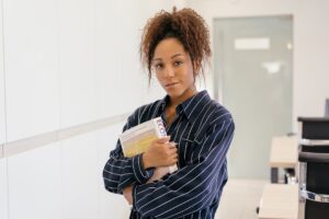 Confident woman with curly hair and striped shirt holding books indoors.