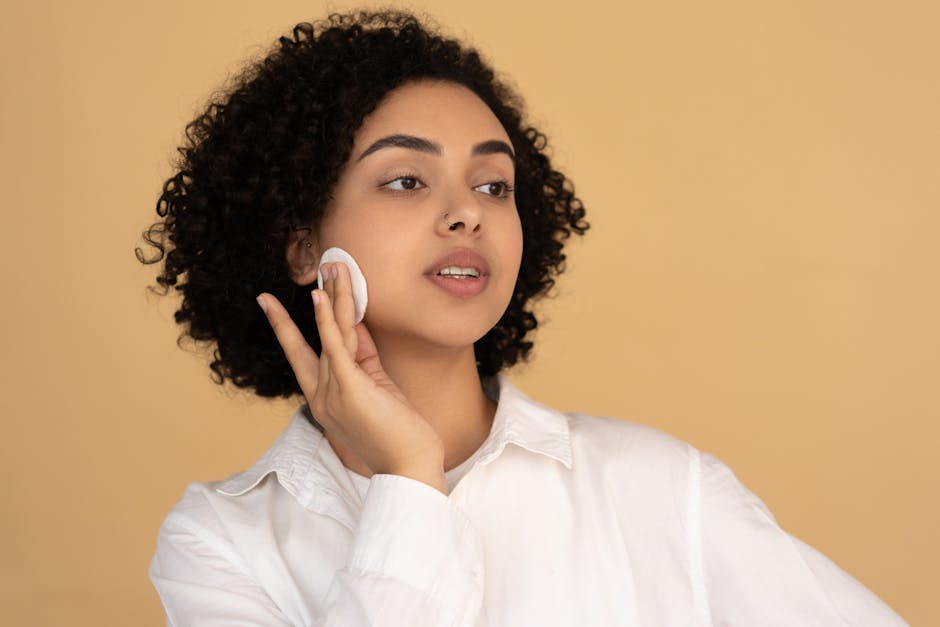 A young woman with curly hair applies skincare product using a cotton pad, enhancing her healthy complexion.