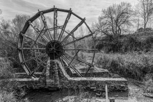 Black and white photo of a rustic water wheel in a serene winter landscape.