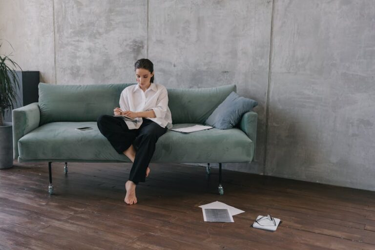 Woman sitting on a couch in an industrial-style living room, engaged in reading or working.