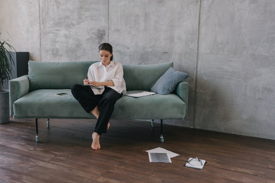 Woman sitting on a couch in an industrial-style living room, engaged in reading or working.