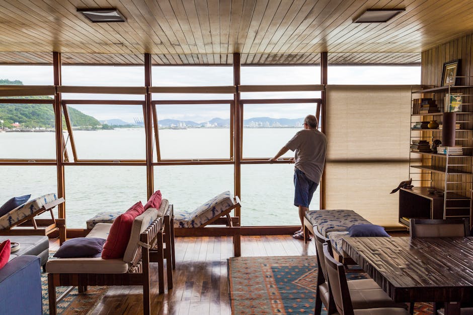 An elderly man stands indoors, gazing out a large window with a view of a scenic bay and mountains in Brazil.