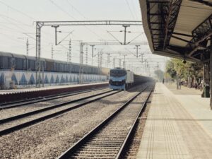 A scenic view of a train approaching an empty railway station platform under a sunny sky.