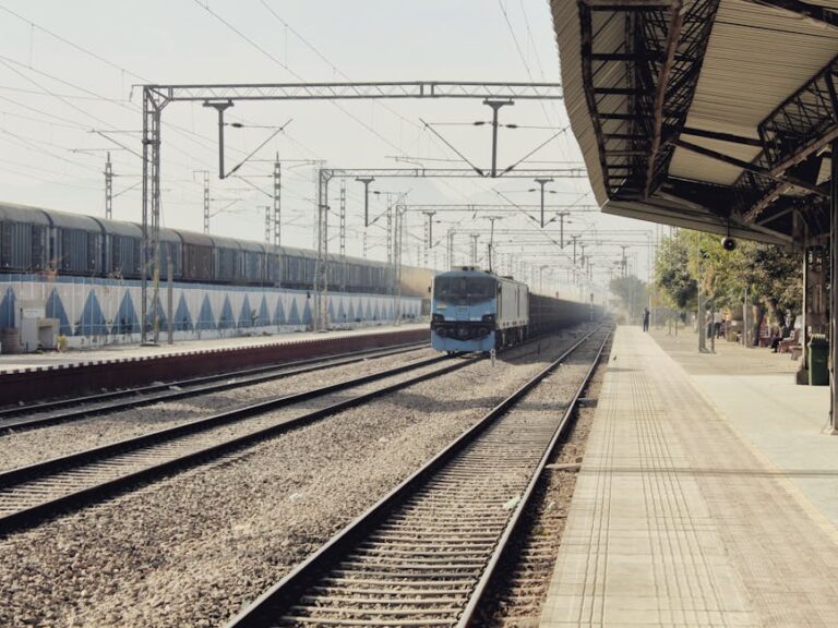 A scenic view of a train approaching an empty railway station platform under a sunny sky.