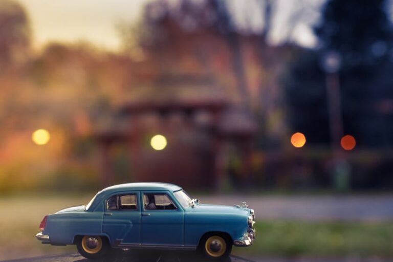 A vintage blue toy car with a blurred city background, photographed outdoors at twilight.