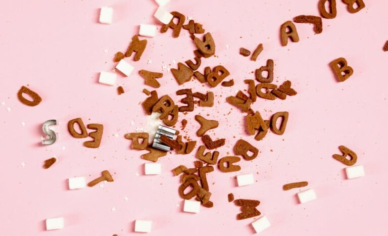 Overhead view of sugar cubes and cookie letters scattered on a pink surface, suggesting sweet chaos.