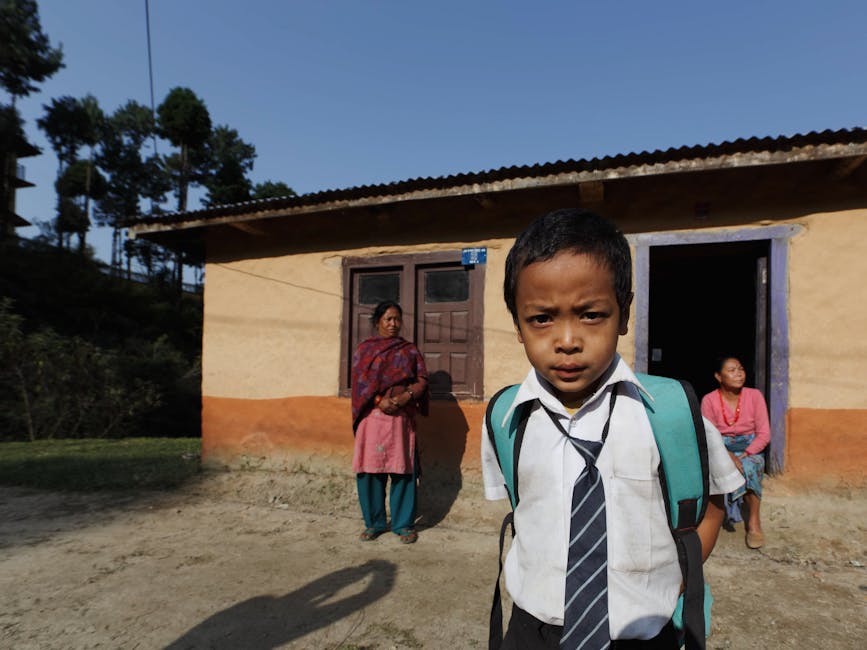 A young boy in school uniform stands in front of a rural house with two women in the background.