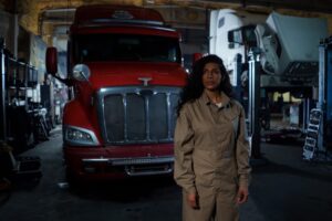 A female auto mechanic stands confidently in a dimly lit repair shop with trucks in the background.