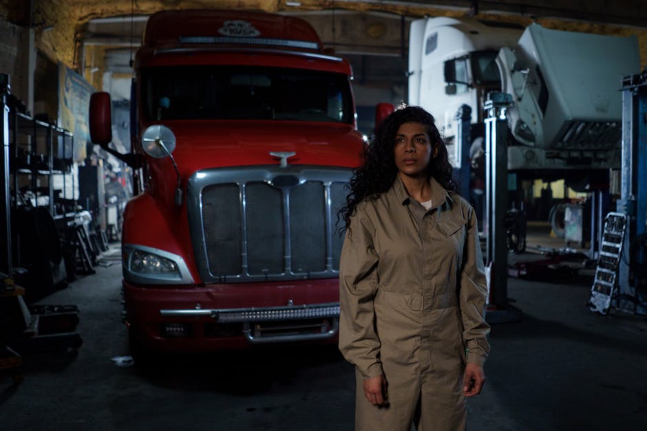 A female auto mechanic stands confidently in a dimly lit repair shop with trucks in the background.