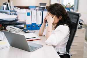 A woman in an office sitting at a desk, visibly stressed, using a phone. Workplace stress concept.