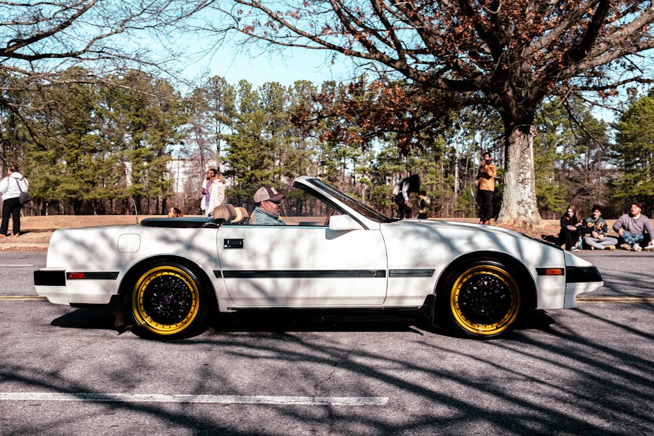 A vintage white convertible with yellow wheels on a park road under a clear sky.