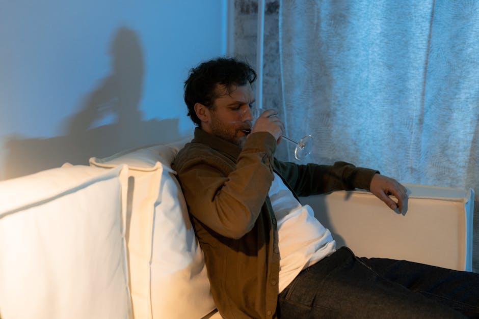 A man sits on a couch, drinking wine in a dimly lit room, conveying a contemplative mood.