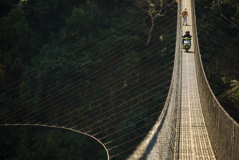 Person riding a scooter on a suspension bridge through dense forest.