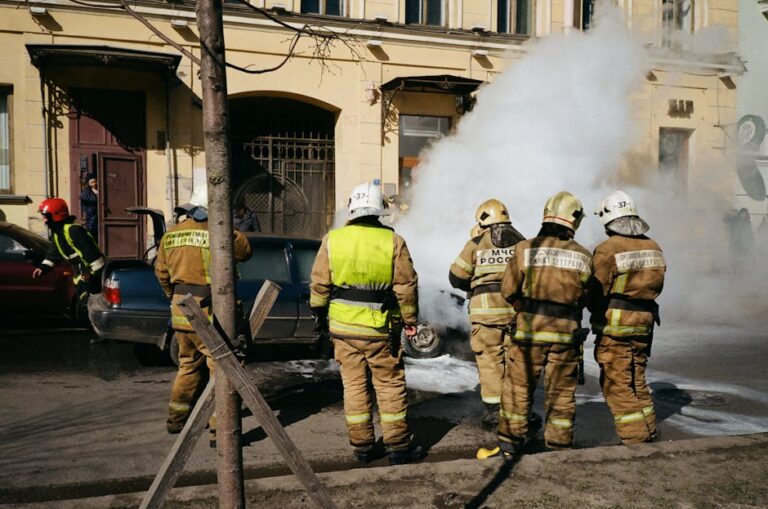 Firefighters responding to a car fire on a street in St. Petersburg, Russia, with smoke and safety gear in view.